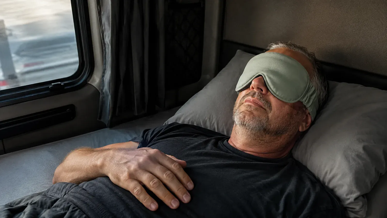 A man lying on his back in a truck bunk wearing a sage green weighted sleep mask with contoured eye cups