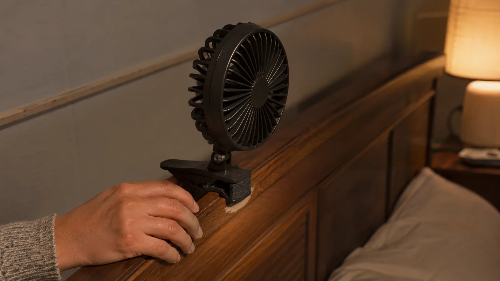 Close-up of a hand clamping the BESKAR fan onto the wooden edge of a bedroom headboard, the fan's clamp and blade guard visible at a new angle