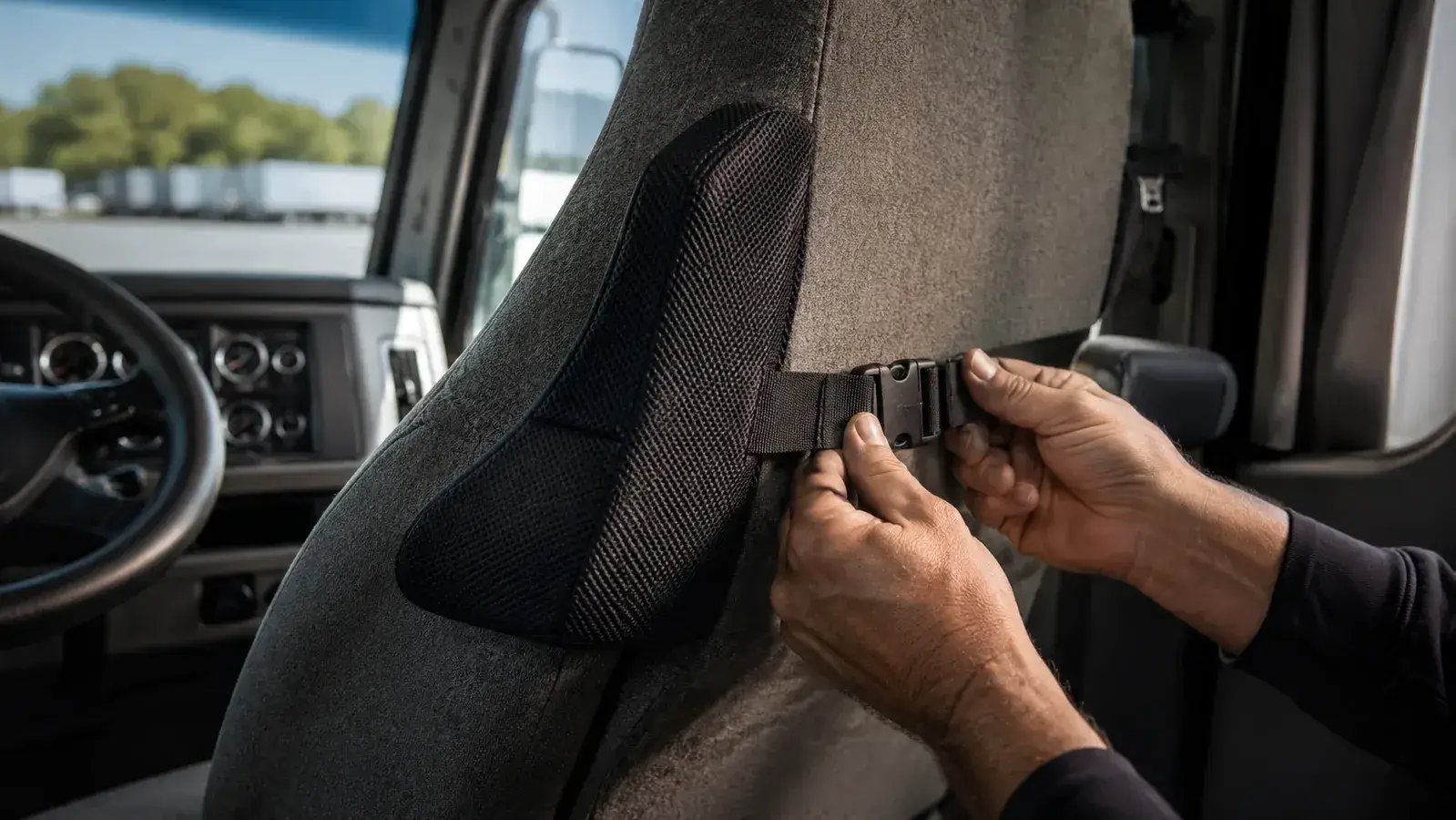 Close-up of a man's hands buckling the elastic strap of a black lumbar pillow around the back of a cloth truck seat