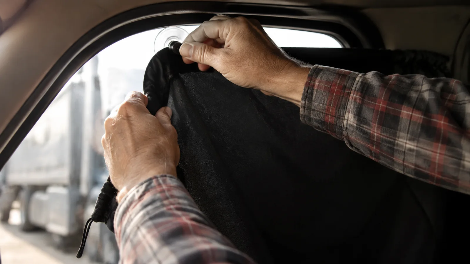 A driver's hands pressing a suction cup into the corner of a black portable blackout curtain against a curved truck cab side window