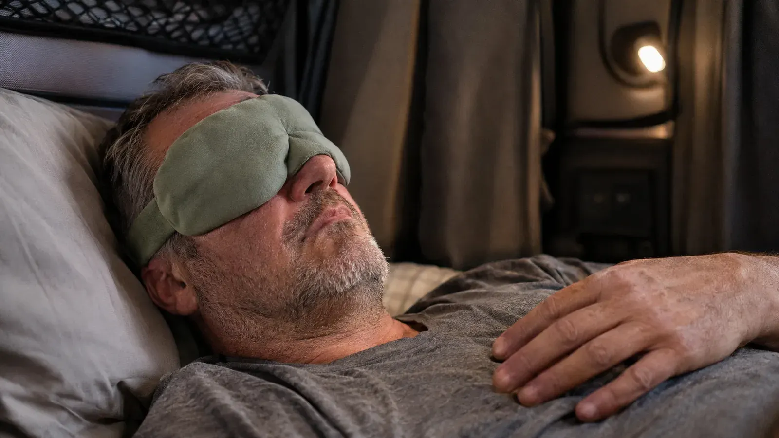 A truck driver lying in a sleeper cab bunk wearing a sage green nodpod weighted eye mask, curtain visible behind him