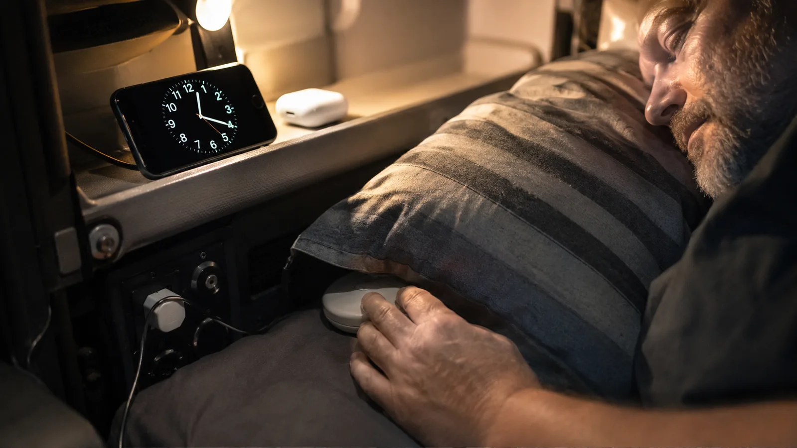 A trucker's nightstand shelf in a sleeper cab showing a phone propped on a vibrating alarm clock stand next to a small earbud charging case