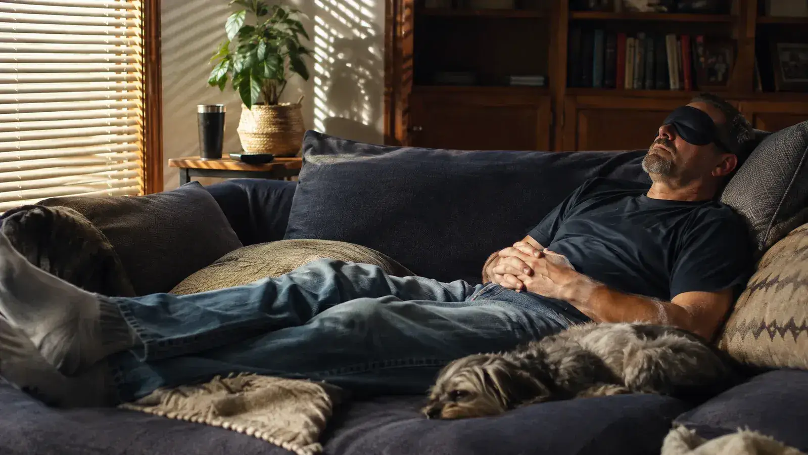 A man napping on a home couch wearing a sleep mask with warm afternoon light coming through the blinds.