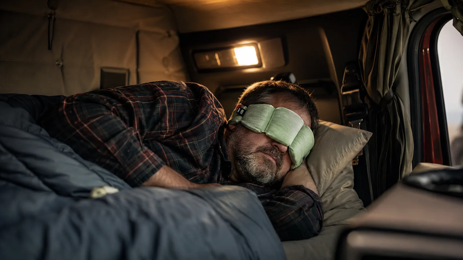 A man lying in a truck bunk wearing a sage green nodpod weighted sleep mask, finally asleep despite light outside