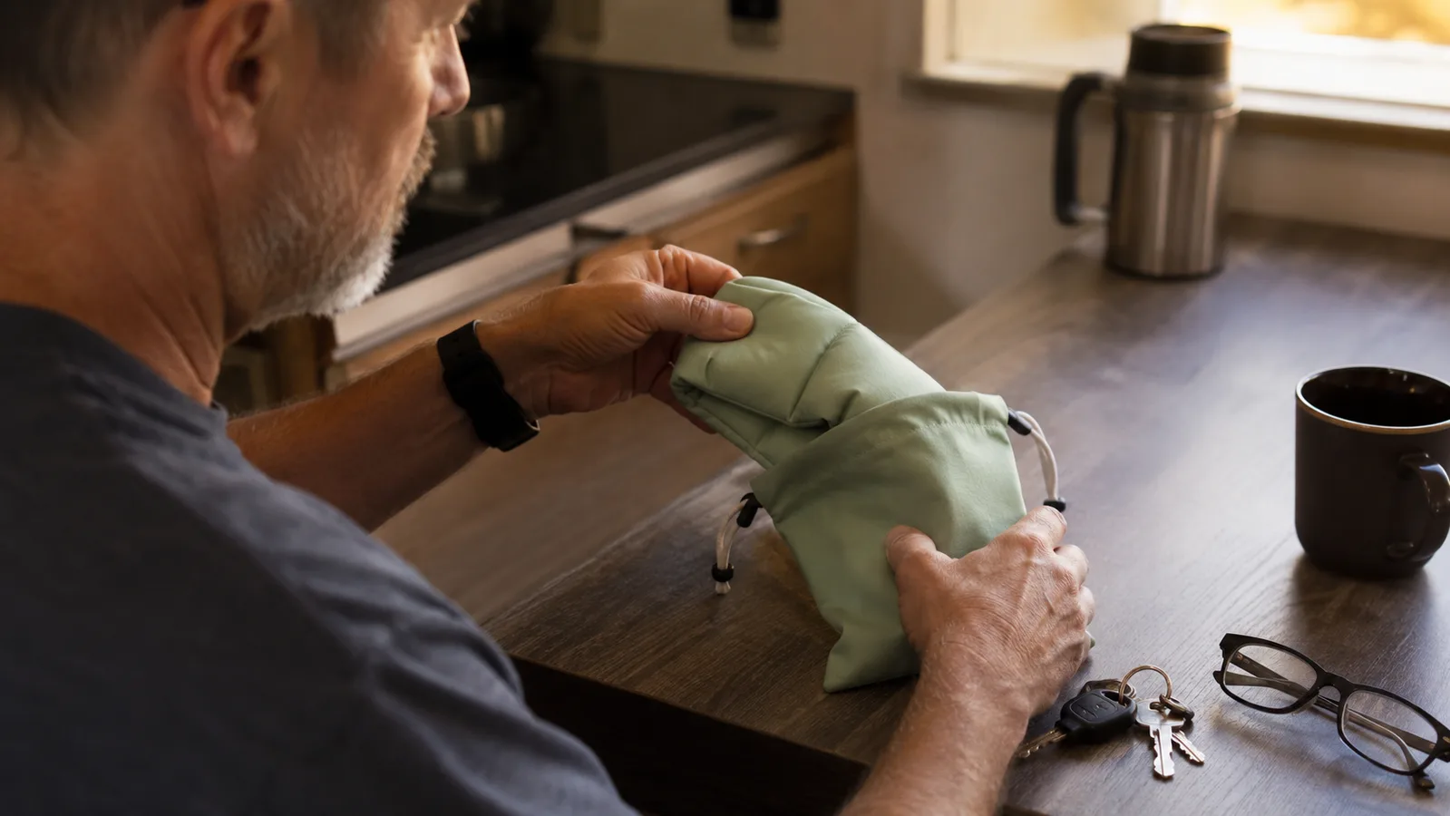 Man folding a sage green weighted sleep mask into a small zip pouch on a kitchen counter at home