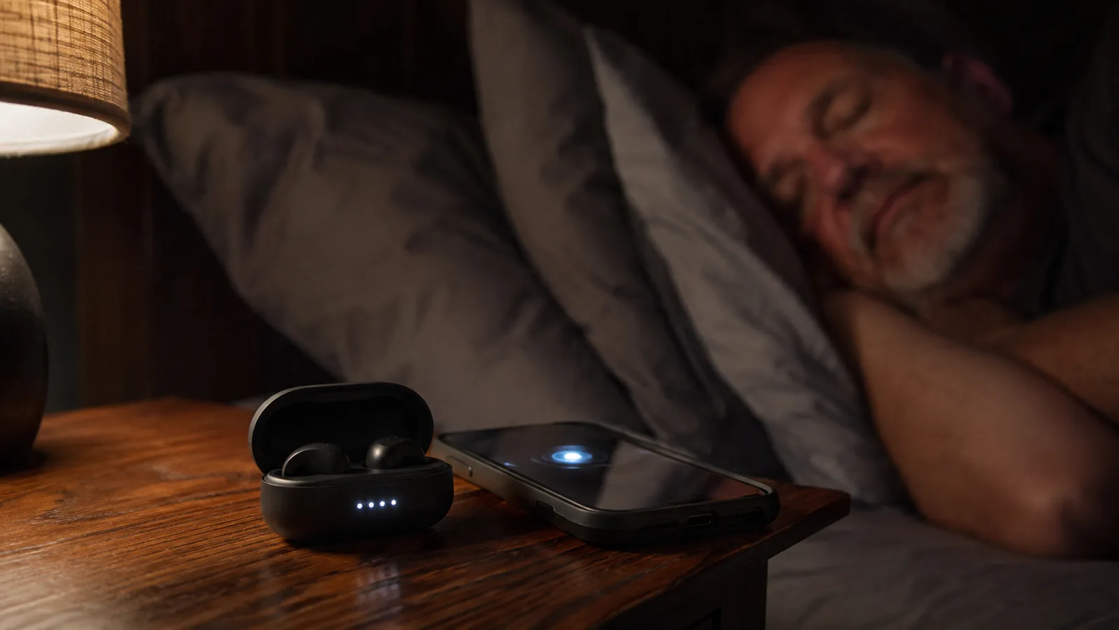 The small charging case for sleep earbuds sitting on a nightstand next to a phone with the alarm screen lit up